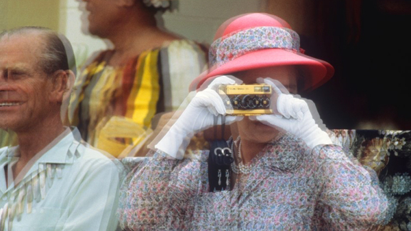 A photography taken on the move: on the left there's an old man, with a white shirt, smiling; behind him, a body with a vivid yellow dress. In the center, an old woman with white gloves and a red hat taking a picture with a yellow camera